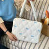 A white quilted laptop shoulder bag with blue bows is resting on a striped surface next to a person's arm.