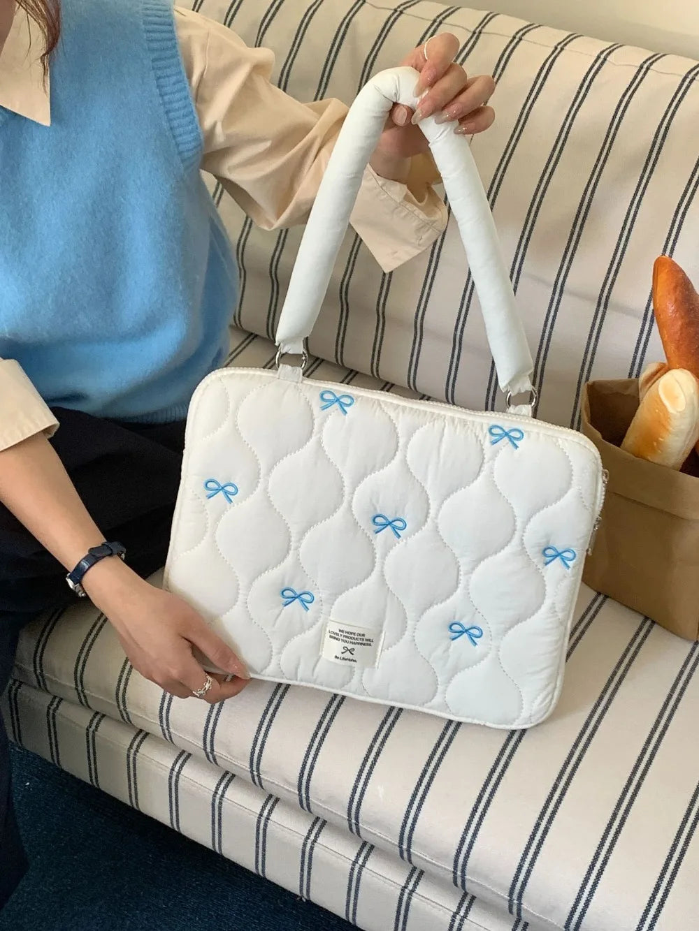 A white quilted laptop shoulder bag with blue bows is resting on a striped surface next to a person's arm.