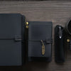 A black handcrafted leather refillable journal next to a leather pen and a mug on a dark wooden surface.