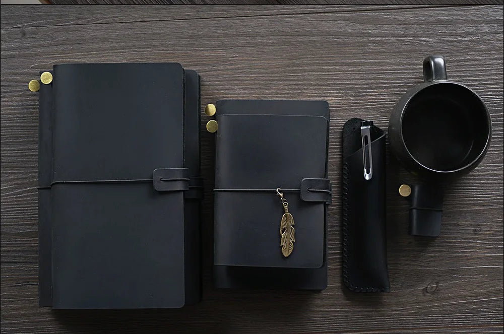 A black handcrafted leather refillable journal next to a leather pen and a mug on a dark wooden surface.
