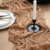 A beige crinkle gauze table runner draped across a wooden surface with a white candle in a black holder.