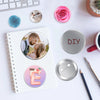 A DIY pin back button kit featuring a blank silver button and paper inserts, surrounded by craft supplies.