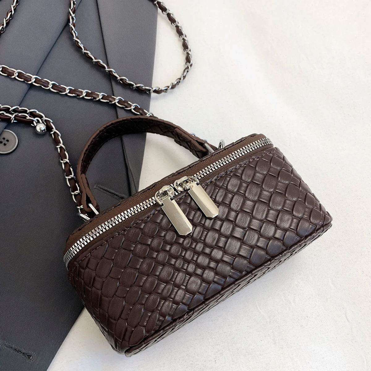 A dark brown woven PU leather crossbody bag with a silver zipper and chain resting on a gray envelope.