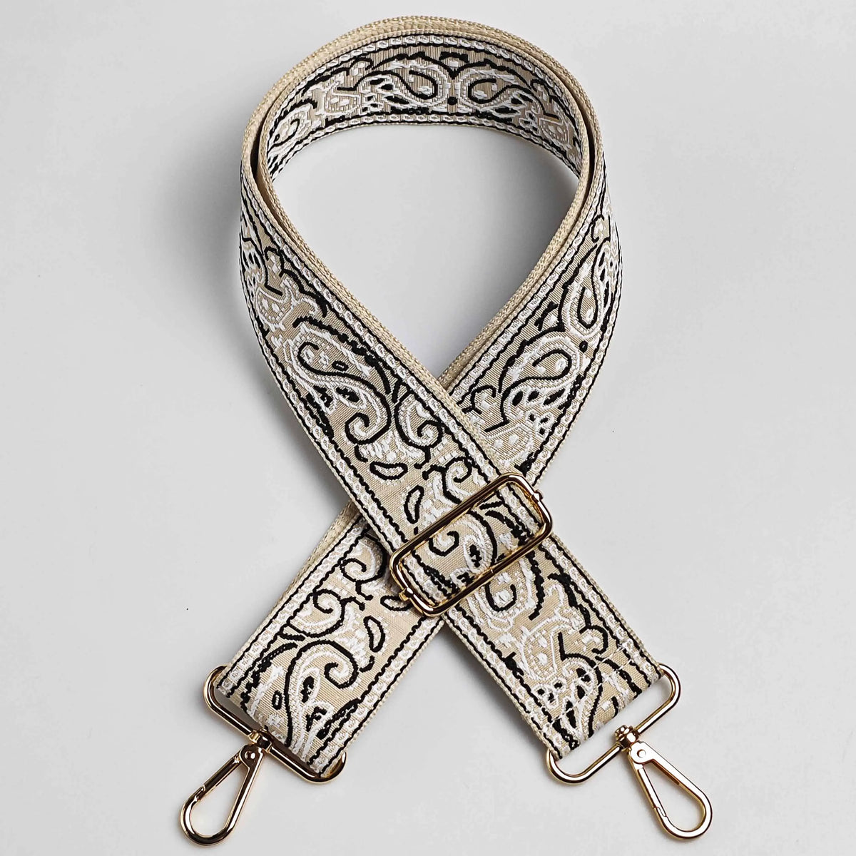 A beige and black paisley jacquard strap for a bag, featuring an X-shaped design and gold hardware.