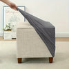 A hand is draping a gray stretch jacquard ottoman slipcover over a beige upholstered ottoman.