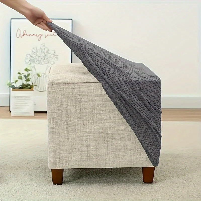 A hand is draping a gray stretch jacquard ottoman slipcover over a beige upholstered ottoman.
