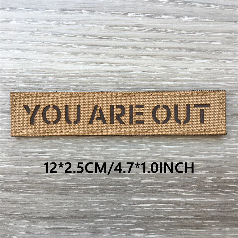 A brown woven patch with the text YOU ARE OUT on it, measuring 12x2.5cm or 4.7x1.0 inch.