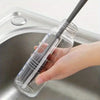 A hand is holding a clear silicone bottle brush with a long handle inside a stainless steel sink.