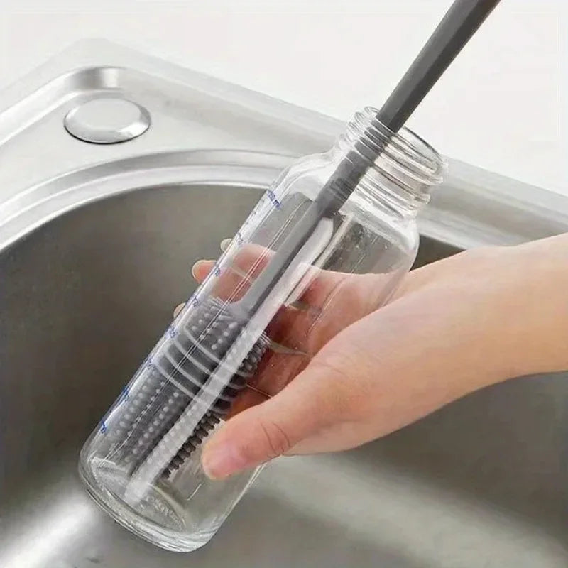 A hand is holding a clear silicone bottle brush with a long handle inside a stainless steel sink.