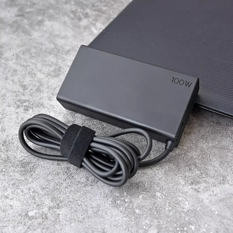 Schwarzer 100W USB-C PD Laptop-Ladegerät mit gewickeltem Kabel auf grauer Oberfläche.