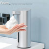 A hand is dispensing foam soap from a stainless steel automatic dispenser onto a white sponge.