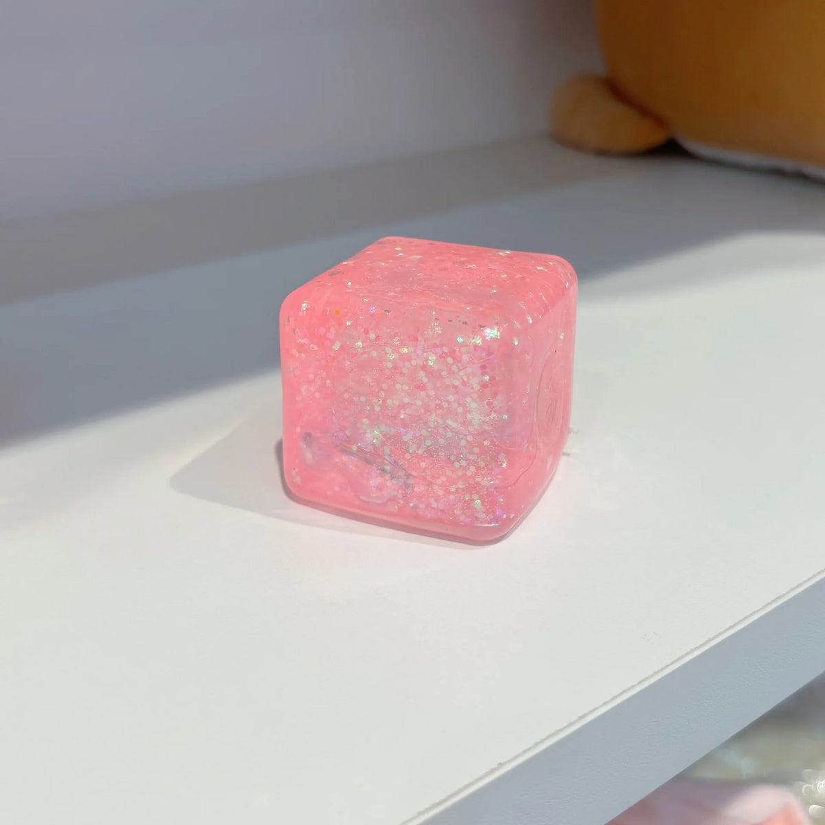 Pinkes, glitzerndes Squishy Spielzeug in Form eines Quadrats auf einer weißen Oberfläche.