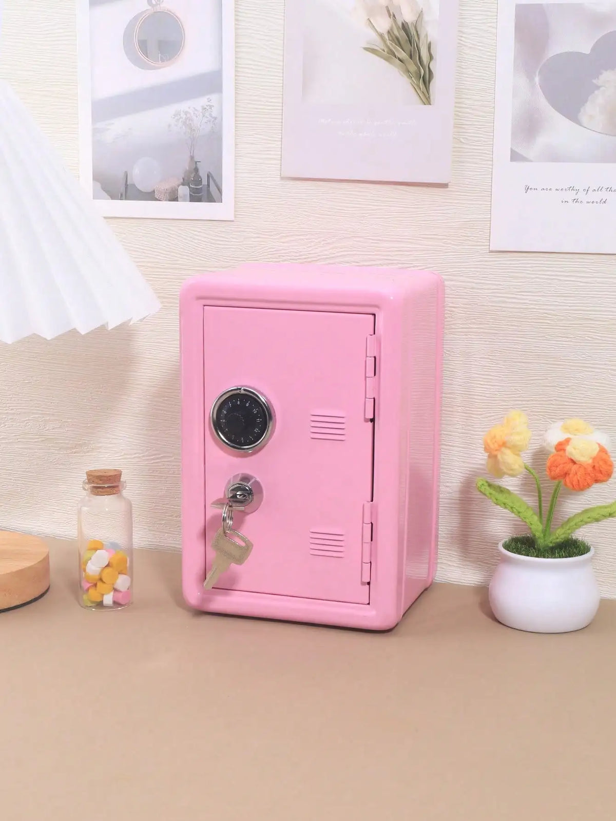 A pink compact desktop safe with a combination lock sits on a light brown surface next to a small glass jar and a potted plan
