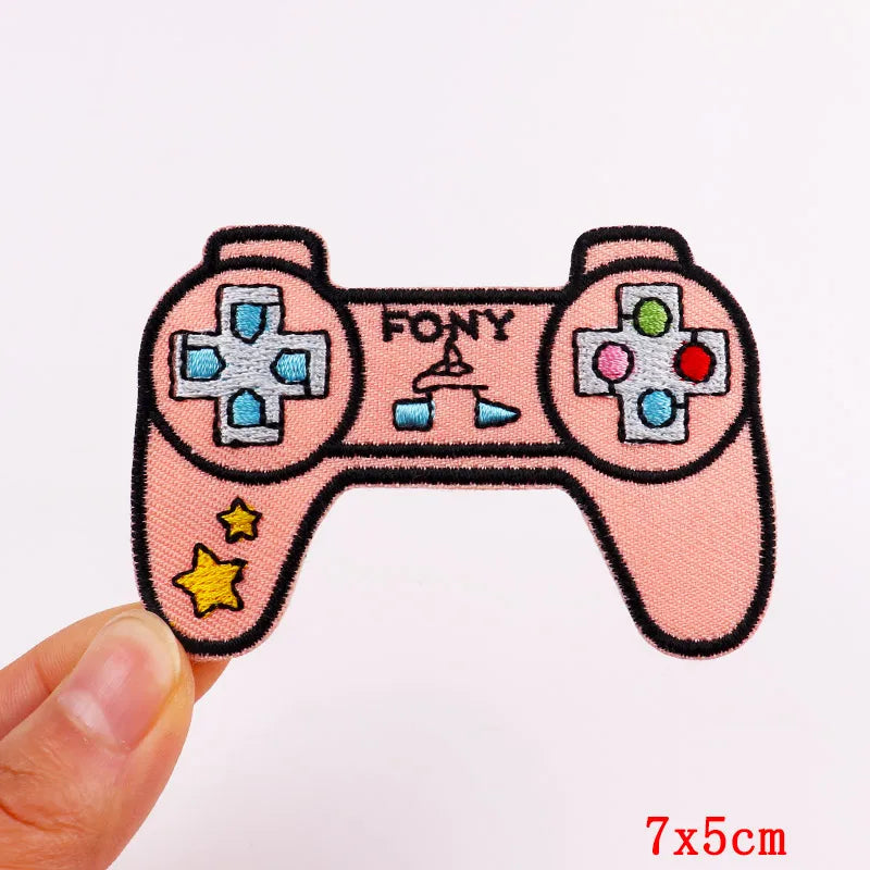 A hand holds a pink embroidered iron-on patch set featuring a retro game controller design.