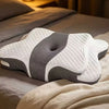 A white and gray ergonomic butterfly memory foam pillow resting on a bed with soft bedding.
