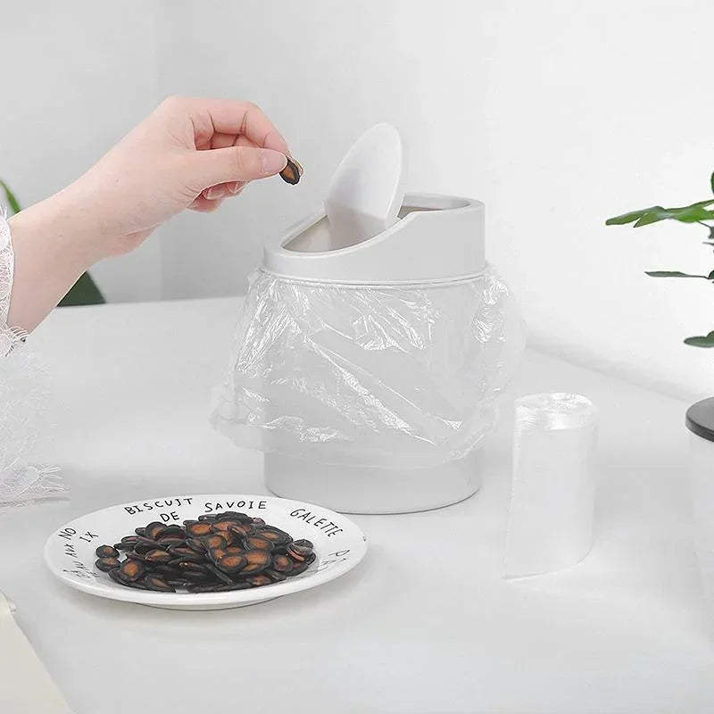 A hand is using a flip lid to access the interior of a compact covered desktop trash bin.