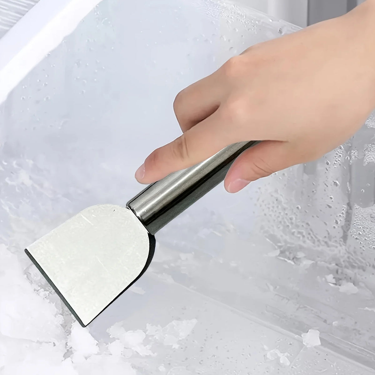 A hand is using a stainless steel multi-purpose scraper to remove ice and snow from a window pane.