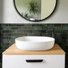 A white vessel sink is displayed on a wooden countertop above a dark subway tile backsplash and a round mirror.