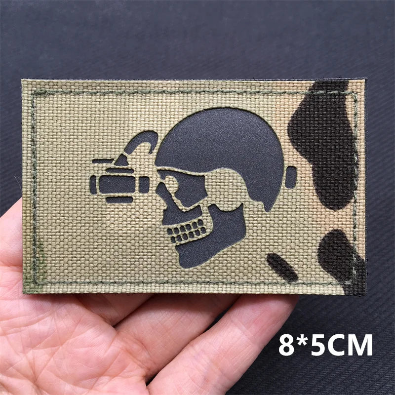 A rectangular tactical patch featuring a silhouette of a skull and crossbones with a camera, made of olive green material.