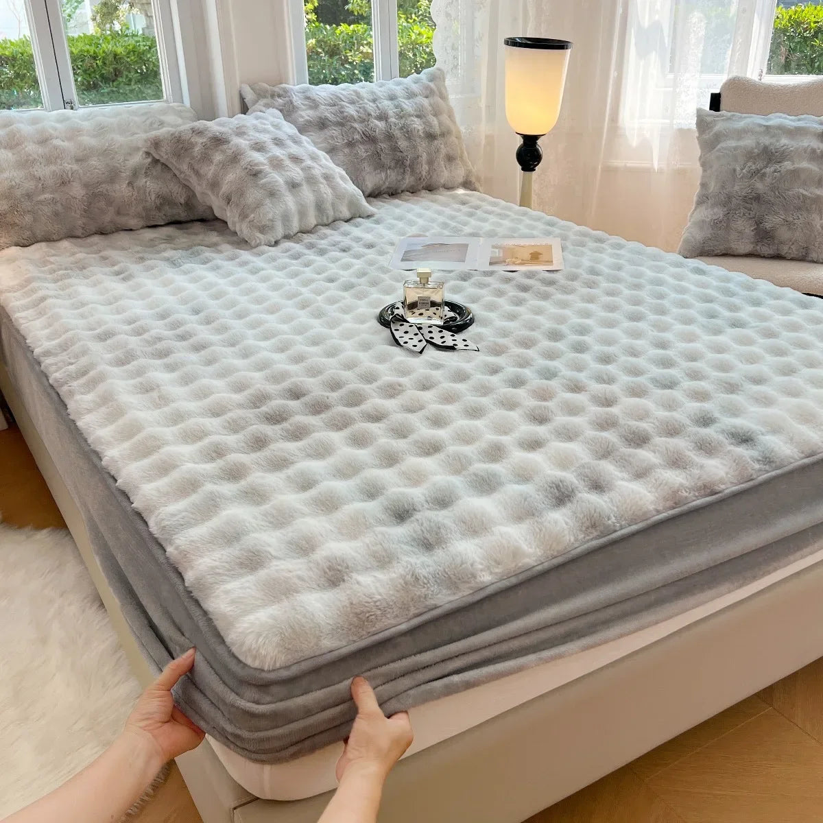 A luxury faux fur fitted sheet and pillowcase set displayed on a bed with sheer curtains and a lamp.