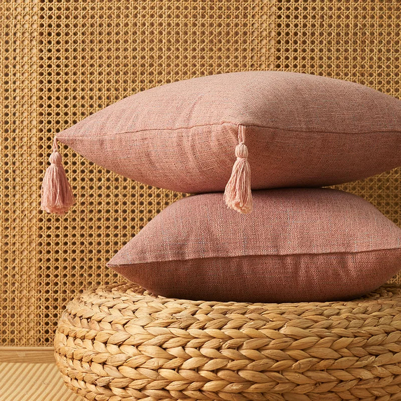 Two dusty rose cotton linen throw pillows with tassels resting on a woven natural fiber surface against a textured gold backg