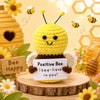 A yellow crochet bee doll with black antennae sits on a wooden slice surrounded by flowers and honeycomb elements.