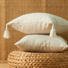 A beige cotton linen throw pillow with tassels rests on a woven natural fiber surface against a textured wall.