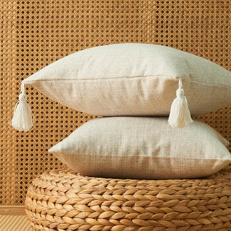 A beige cotton linen throw pillow with tassels rests on a woven natural fiber surface against a textured wall.