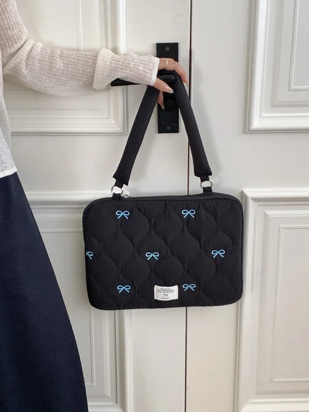 A black quilted laptop shoulder bag with blue bows is hanging on a white door, held by a person's hand.