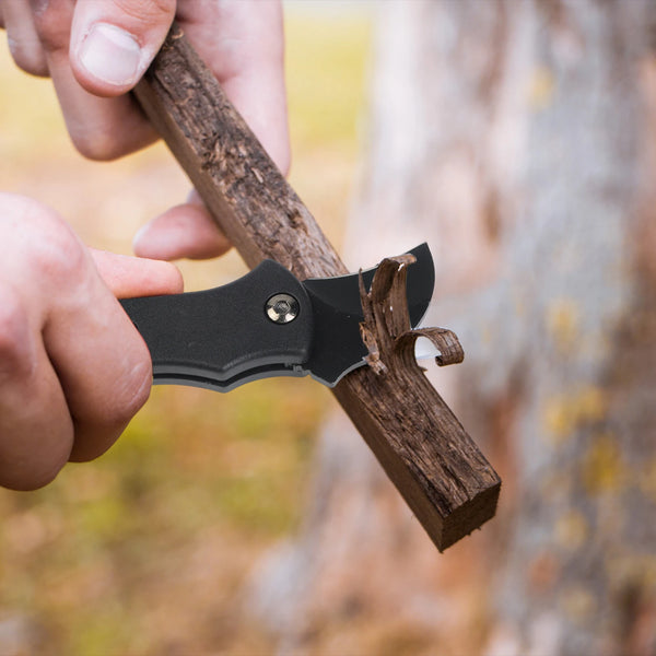A hand holds a black folding utility knife with a wooden handle against a blurred outdoor background.