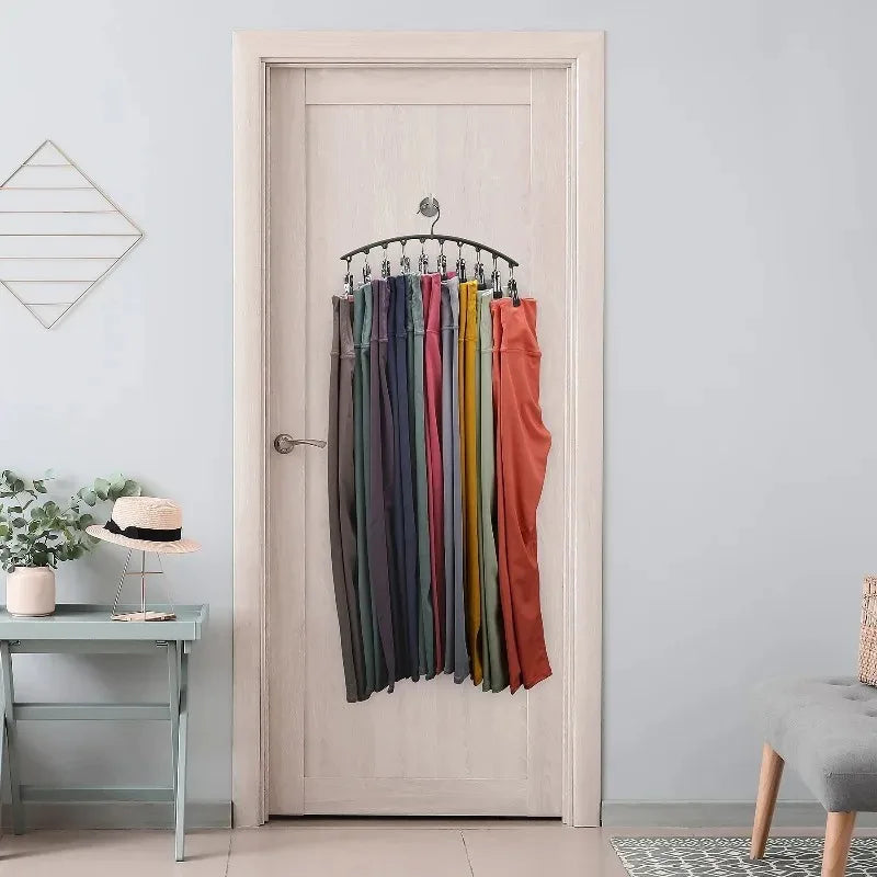 A metal pant hanger with ten clips hanging on a white door, displayed next to a potted plant and a small table.