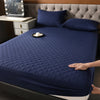 A dark blue quilted waterproof mattress protector is shown on a bed, with pillows and a bedside table visible.
