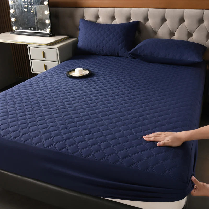 A dark blue quilted waterproof mattress protector is shown on a bed, with pillows and a bedside table visible.