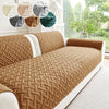 A tan quilted anti-slip sofa cushion is shown on a white sofa, with natural light and greenery in the background.