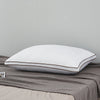 A white luxury goose down and feather pillow with a brown trim resting on a gray fabric.