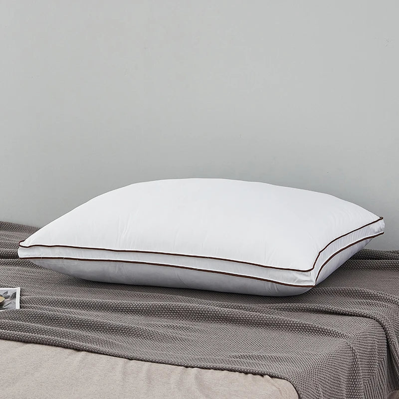 A white luxury goose down and feather pillow with a brown trim resting on a gray fabric.