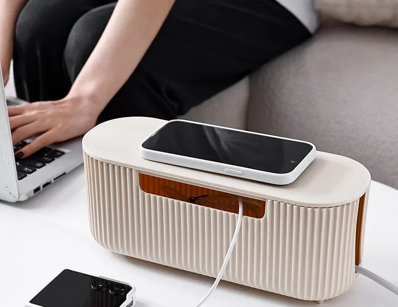 A beige cable management box with a black smartphone resting on it, next to a laptop and power cord.