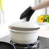 A hand wearing black heat-resistant silicone oven mitts is placing a white ceramic cooking pot onto a stovetop.