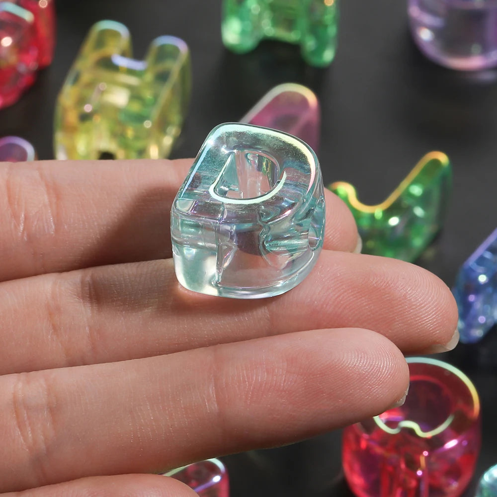 A hand holds a clear acrylic alphabet bead, showing its shape and clarity against a dark background.