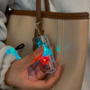A hand holds a clear LED clickable fidget keychain with a keyboard design attached to a leather bag strap.