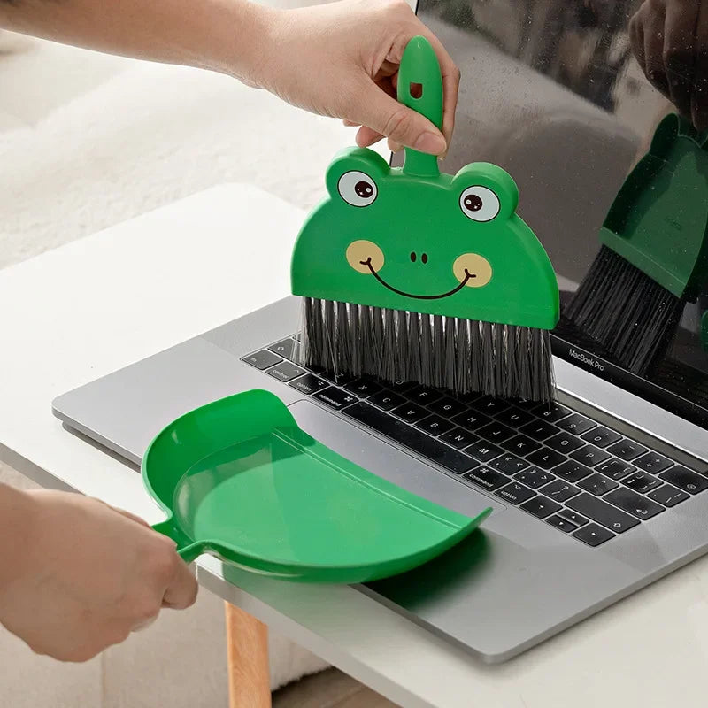 A hand is using a green cartoon broom and dustpan set to clean a laptop surface.