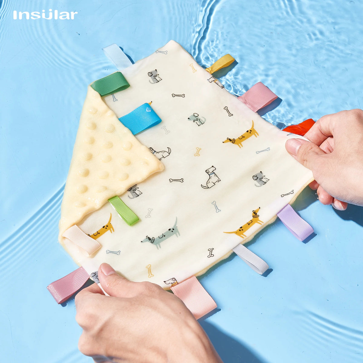 A hand is holding a yellow baby sensory tag comfort blanket with a soothing teether on a light blue surface.