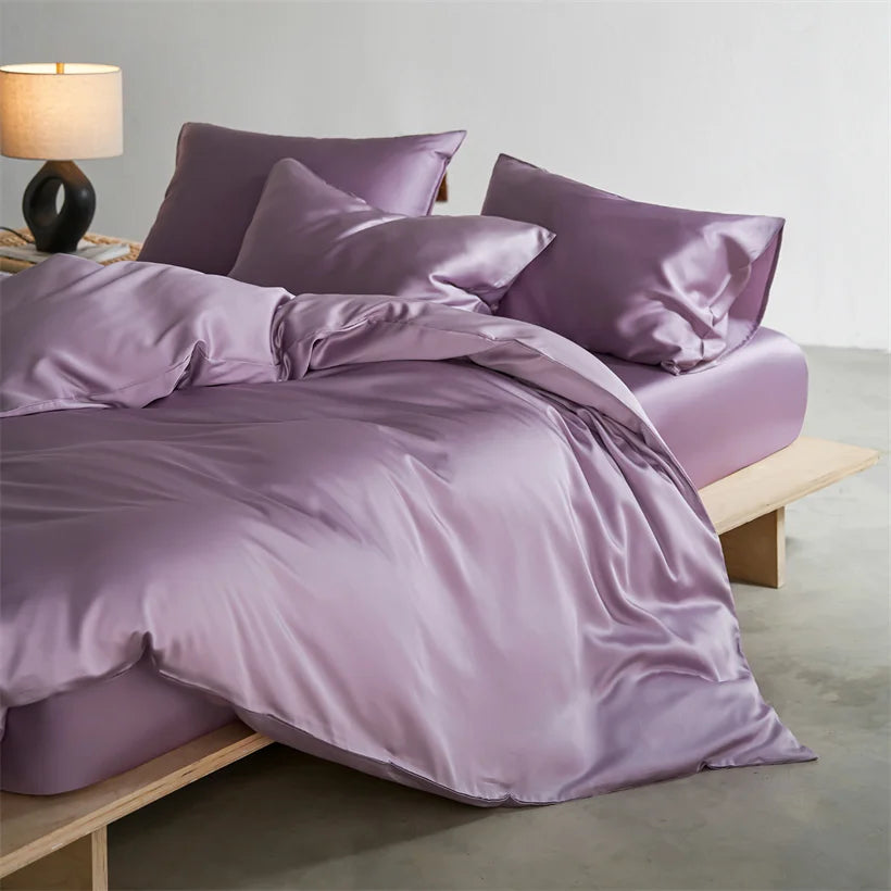 A luxurious lavender silk duvet cover set displayed on a bed with matching pillows and a bedside lamp.
