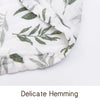 Close-up of a white muslin baby swaddle blanket with green leaf print showing delicate hemming.