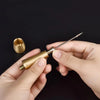 A hand holds a brass leather sewing awl set with a replaceable needle and a separate metal tool on a dark background.