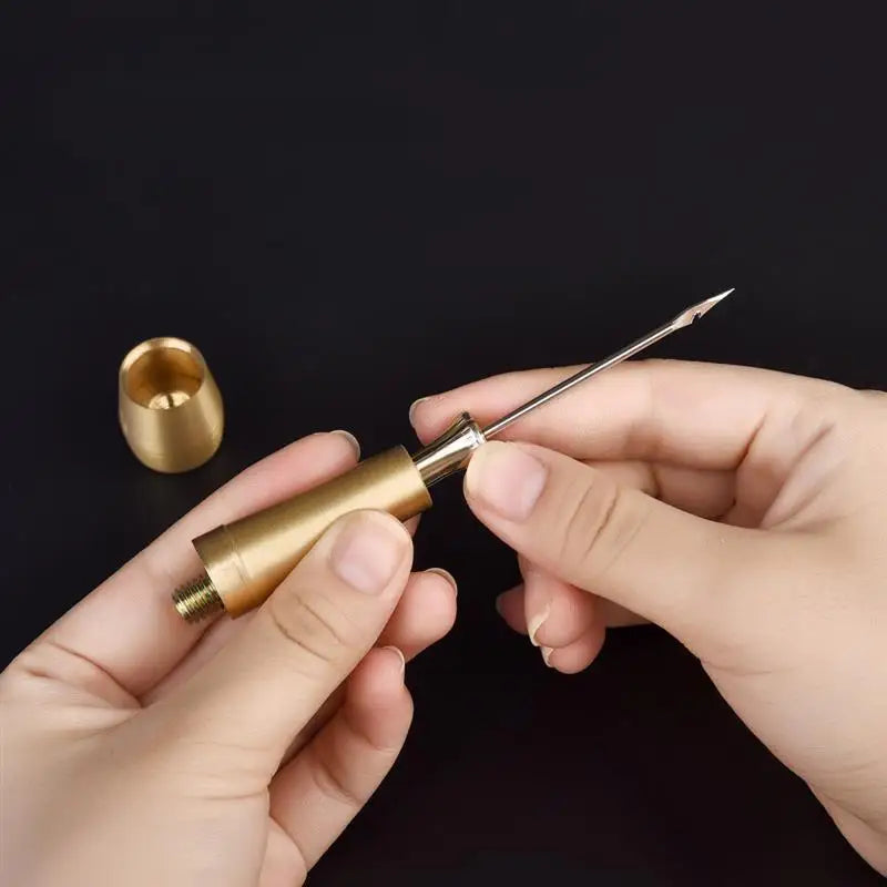 A hand holds a brass leather sewing awl set with a replaceable needle and a separate metal tool on a dark background.