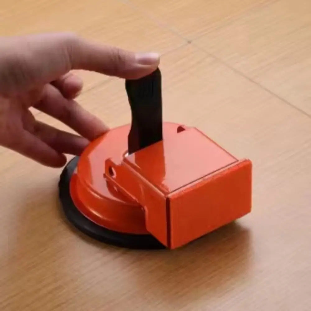 A hand is holding an orange heavy duty floor gap fixer tool with a black suction cup on a wooden surface.