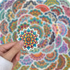 A hand holds a colorful mandala sticker with intricate floral patterns against a patterned background.