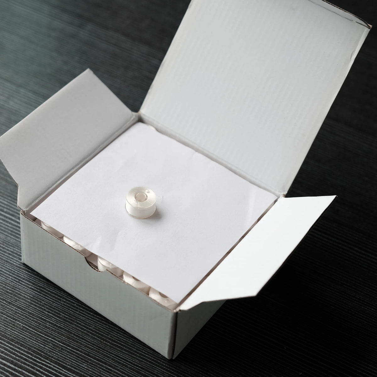 A white box is open, revealing a prewound bobbin with polyester thread inside.