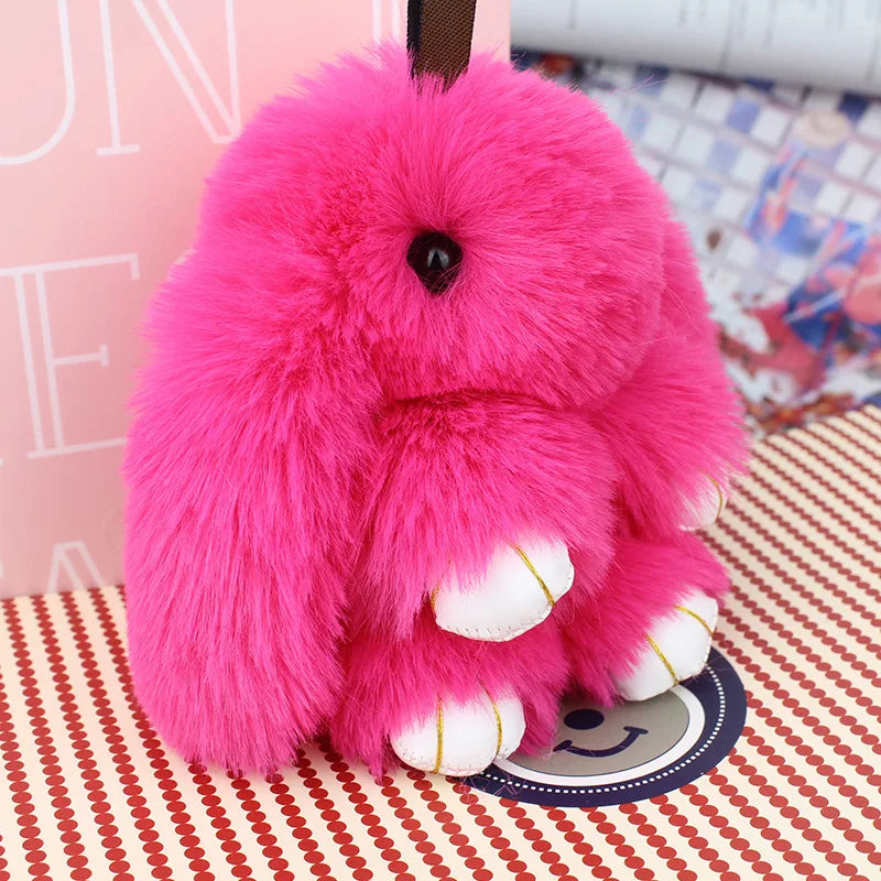 A bright pink fluffy angel bunny keychain with a pom pom tail resting on a patterned surface.